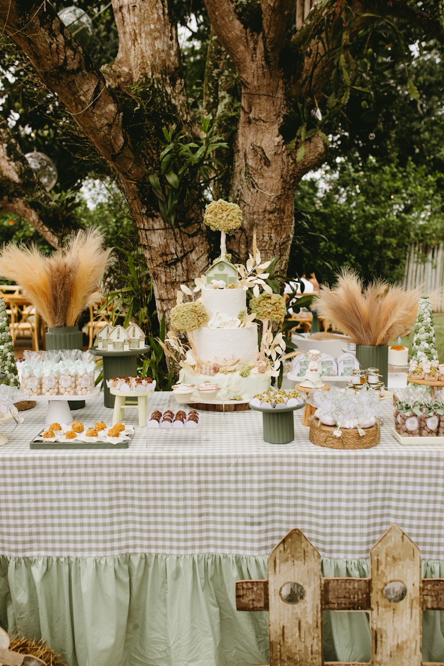 Mesa de doces ao ar livre embaixo de árvore com bolo decorado e arranjos florais